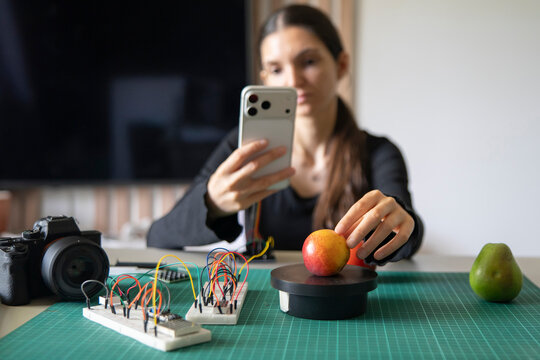 Female educator using smartphone to scan apple on DIY electronic scale for online machine learning course.