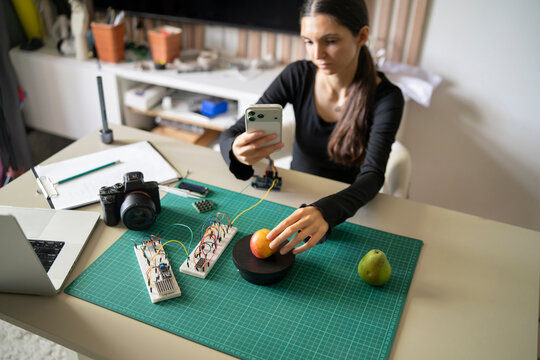 Woman recording STEM tutorial on AI weight scales with smartphone, teaching electronics and coding.