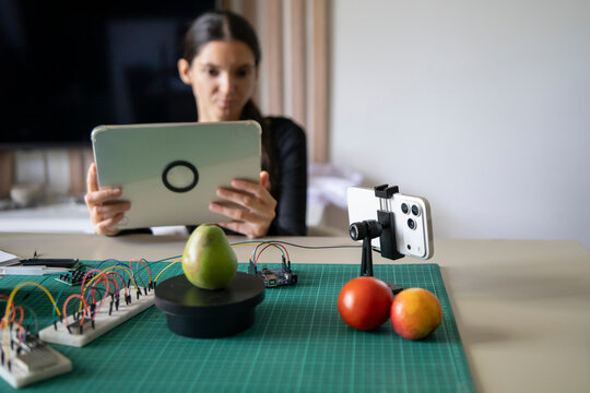 Educator using tablet and smartphone on tripod to record DIY electronic scale tutorial for students.