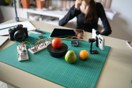 Smart scale with tomato and microcontroller on desk while teacher prepares for online STEM broadcast.