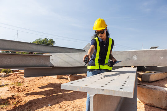 An engineer wearing a yellow hard hat and reflective safety vest inspects structural steel components for a bridge over railway tracks, with steel beams and a drilled connection plate.