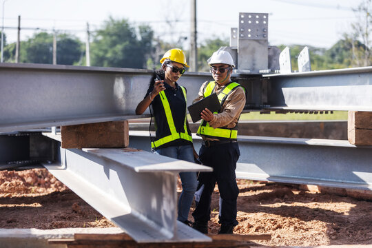 Two engineers wearing hard hats and reflective safety vests inspect structural steel beams at an outdoor construction site, using a tablet and handheld radio to coordinate quality checks.