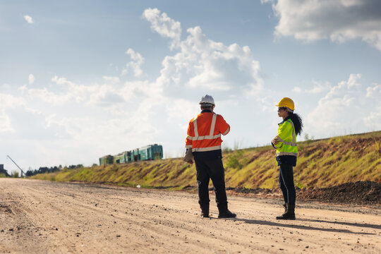 Two engineers wearing hard hats and reflective workwear discuss site planning on unpaved ground at a railway construction project, with a grassy embankment and rail equipment in the background.