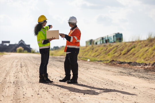Two engineers wearing hard hats and reflective workwear discuss a railway construction project using a laptop on an unpaved worksite, with an embankment and railway machinery in the background.
