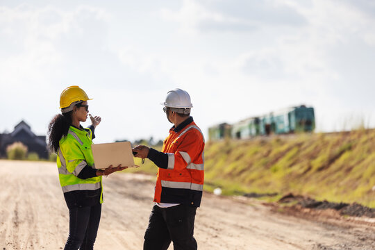 Two engineers wearing hard hats and reflective workwear discuss a railway construction project using a laptop on an unpaved worksite, with an embankment and railway machinery in the background.