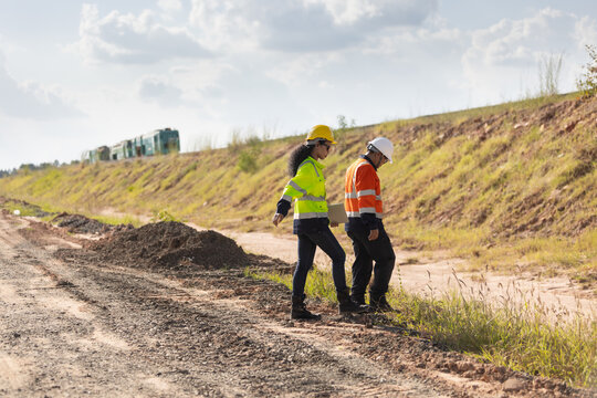 Two engineers wearing hard hats and reflective workwear walk across uneven ground at a railway construction site, inspecting the terrain beside a grassy embankment.