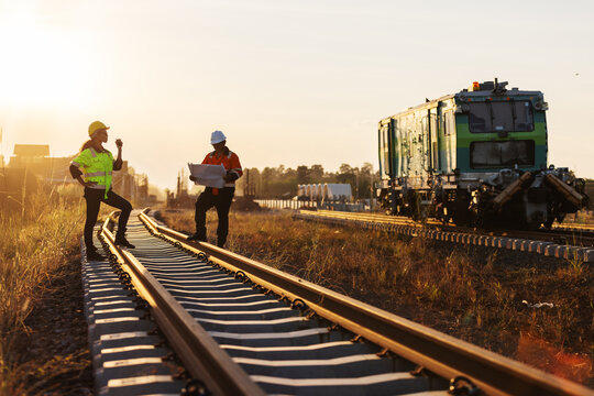 Two engineers in reflective workwear coordinate railway construction in warm evening light. One uses a handheld radio while the other reviews plans
