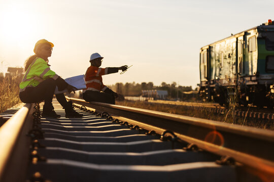 An engineer in reflective workwear and a hard hat reviews documents while seated on a railway rail, with a colleague nearby and rail equipment in the background under warm evening light.