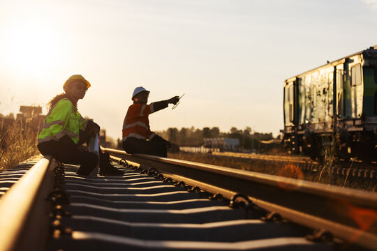An engineer in reflective workwear and a hard hat reviews documents while seated on a railway rail, with a colleague nearby and rail equipment in the background under warm evening light.