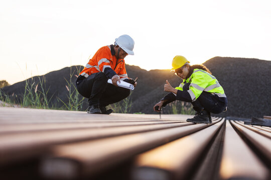 Two engineers in hard hats and reflective workwear inspect steel railway rails at an outdoor site. One checks the rail surface with a handheld tool while the other records findings on a clipboard.