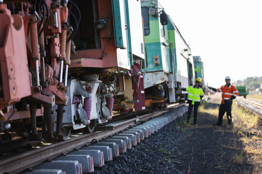 Two engineers in hard hats and reflective workwear inspect mechanical components of railway track construction machinery. One examines the equipment while the other holds a handheld radio.
