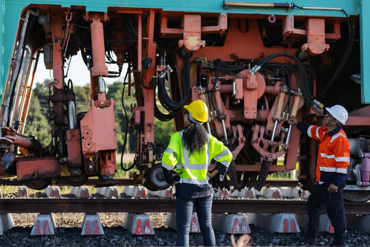 Two engineers in hard hats and reflective workwear inspect mechanical components of railway track construction machinery. 