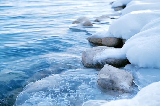 Winter riverbank with stones covered in snow and ice, clear blue water and iced bubbles, peaceful cold nature landscape.