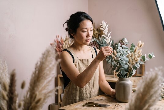 Focused Asian female florist in a linen apron carefully arranging dried flowers and eucalyptus in a ceramic vase at her creative floral studio