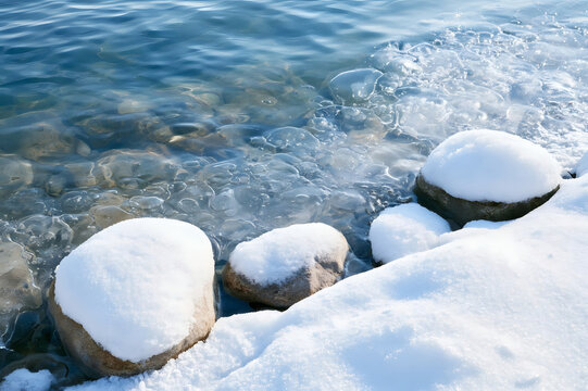 Winter landscape with snow-covered stones on the shore and clear icy water. Tranquil nature scene in cold weather.