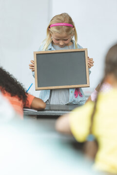 Schoolgirl holds slate in classroom with classmates in front.