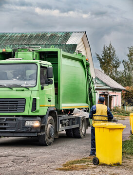 A worker watches as a green garbage truck stops to collect waste in a neighborhood. The scene shows houses and a yellow bin nearby