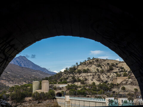 View of arid hills and concrete structures near Amadorio dam framed by dark stone arch.