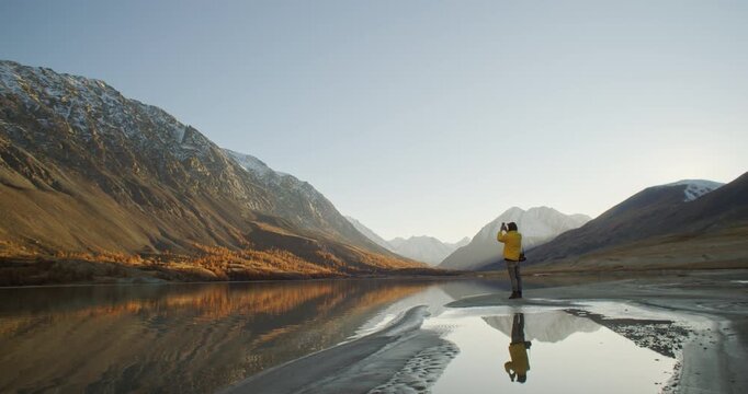 Traveler in a yellow jacket taking a photo with a smartphone while standing by a calm mountain lake at sunrise. Snow covered peaks, soft golden light, and clear reflections create a dramatic outdoor