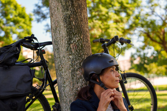 Hispanic woman putting on helmet by tree with bicycle in M&aacute;laga park