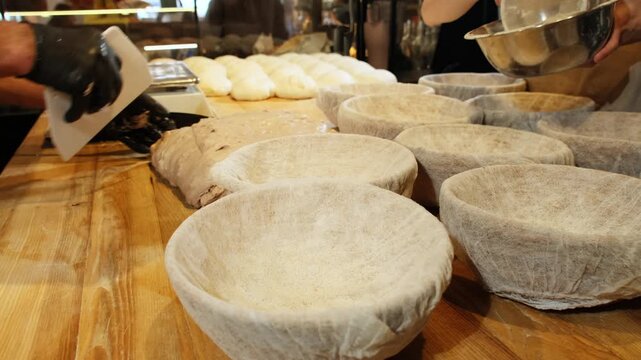 Baker dusts round proofing baskets with flour on wooden bakery table. Skilled worker shakes fine flour over liners preparing baskets for dough