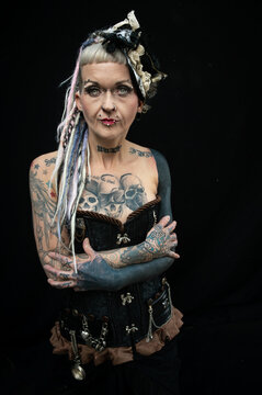 Tattooed woman in steampunk corset with dreadlocks and body piercings posing against black background