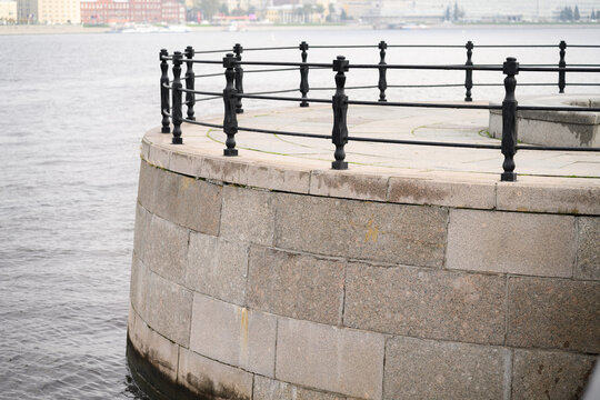Granite embankment parapet with black metal railing over water, city on background, overcast day.