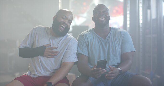 African American men laughing while looking at smartphone on gym bench with wrist support and watch