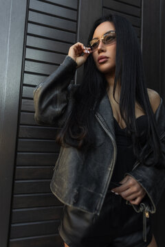 Stylish Asian woman in a leather jacket and sunglasses looking up thoughtfully, posing from a low angle against a dark textured background.