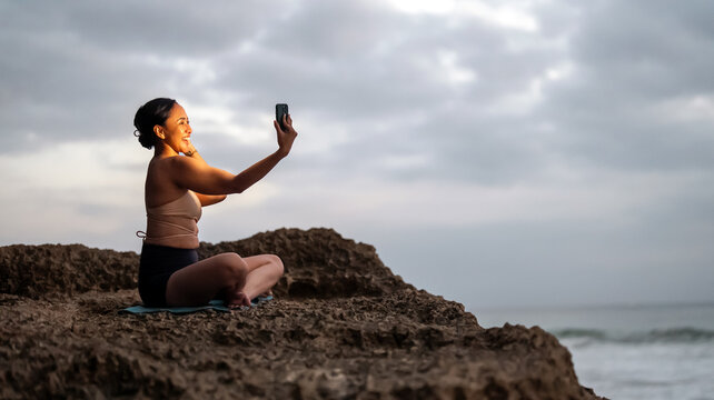 Asian woman taking a smartphone selfie while sitting in yoga pose on an ocean cliff