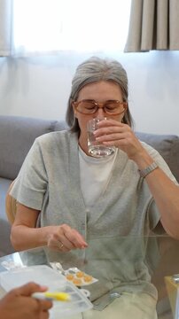 Senior woman taking prescribed oral medicine with a glass of water while sitting next to home health nurse in living room