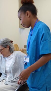 Hispanic female nurse assisting an elderly woman to stand up from her wheelchair and sit on the sofa at home