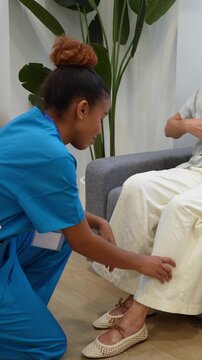 Attentive latina home health nurse kneeling while examining the leg and ankle of an elderly female patient sitting on a couch
