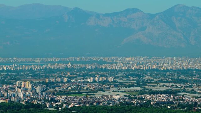Aerial tracking shot following a passenger airplane approaching and landing at Antalya Airport in Turkey, with airport infrastructure, urban Antalya and distant Taurus Mountains across the horizon