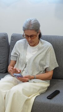 Elderly woman sitting on a sofa in the living room, listening to a voice message and talking on the smartphone