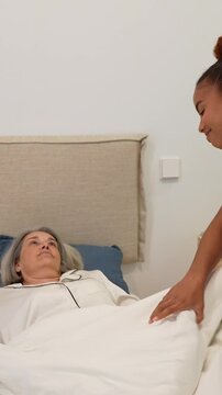 Compassionate latina nurse covering senior woman with blanket and checking her temperature in bedroom, offering home healthcare