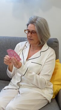 Senior woman wearing pajamas sitting on sofa and holding playing cards while enjoying leisure time at home