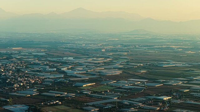 Wide aerial panorama over vast greenhouse farms near Antalya, Turkey, with hundreds of plastic-covered agricultural structures, cultivated fields, farm roads and hazy Taurus Mountains on the horizon