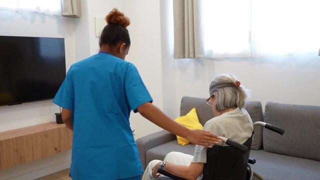 Compassionate female caregiver assisting an elderly disabled woman to stand up from a wheelchair and sit on the couch indoors