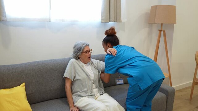 Attentive latina home health nurse assisting an elderly woman with back pain to stand up from the living room sofa