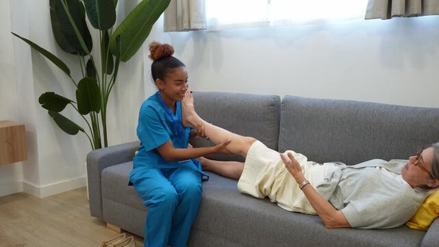 Latina home health nurse helping senior woman lying on sofa with leg rehabilitation exercises during medical visit