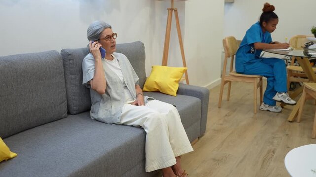 Senior woman talking on mobile phone while sitting on living room sofa as latina home health nurse writes notes at table