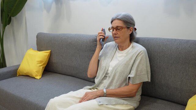 Senior grey haired woman wearing glasses sitting on a modern couch while making a phone call with her smartphone inside living room
