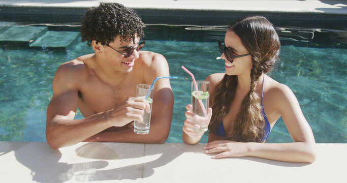 Happy diverse couple in sunglasses standing in sunny swimming pool with drinks