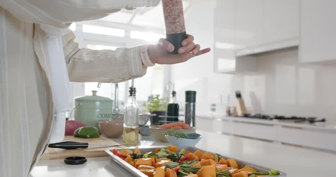 Home cook grinding pink salt over metal tray of cubed vegetables on white kitchen counter
