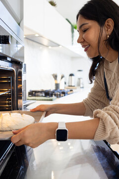 Asian woman holding baking tray and guiding round dough toward open stainless steel oven in kitchen