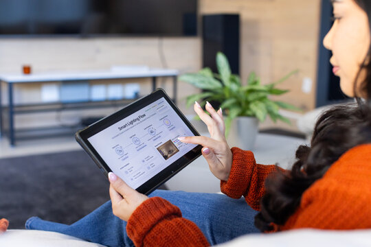 Asian woman in rust-orange sweater sitting on sofa holding tablet pointing at lighting flow diagram