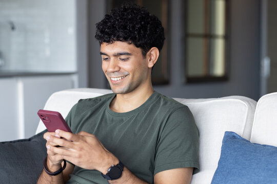 African American man in green t-shirt on white couch using maroon smartphone while smiling