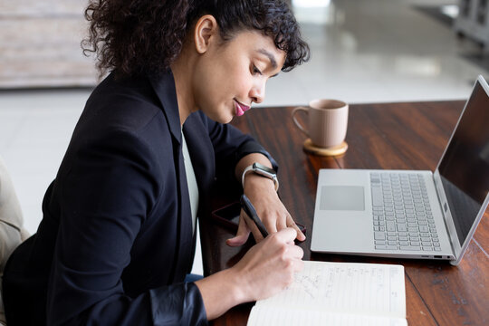 African American woman in black blazer writing in notebook beside open laptop at dark wooden table