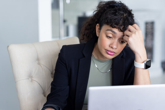 African American woman in business attire sitting at desk resting hand on forehead near laptop